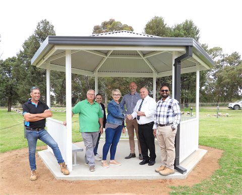 A photo 7 people standing in a white hexagonal gazebo with a dark grey roof. A smiling woman hands a cheque to a man in a white shirt who is shaking her hand.