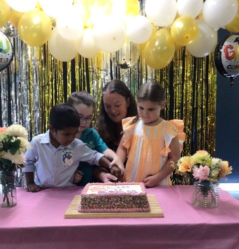 Woman and 3 children hold knife to cut large rectangular cake with sprinkles around it. tinsel and balloons form a backdrop