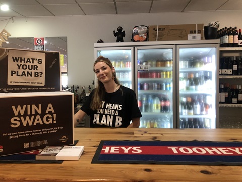 Girl in black Plan B T-shirt standing behind bar with drin fridges in background